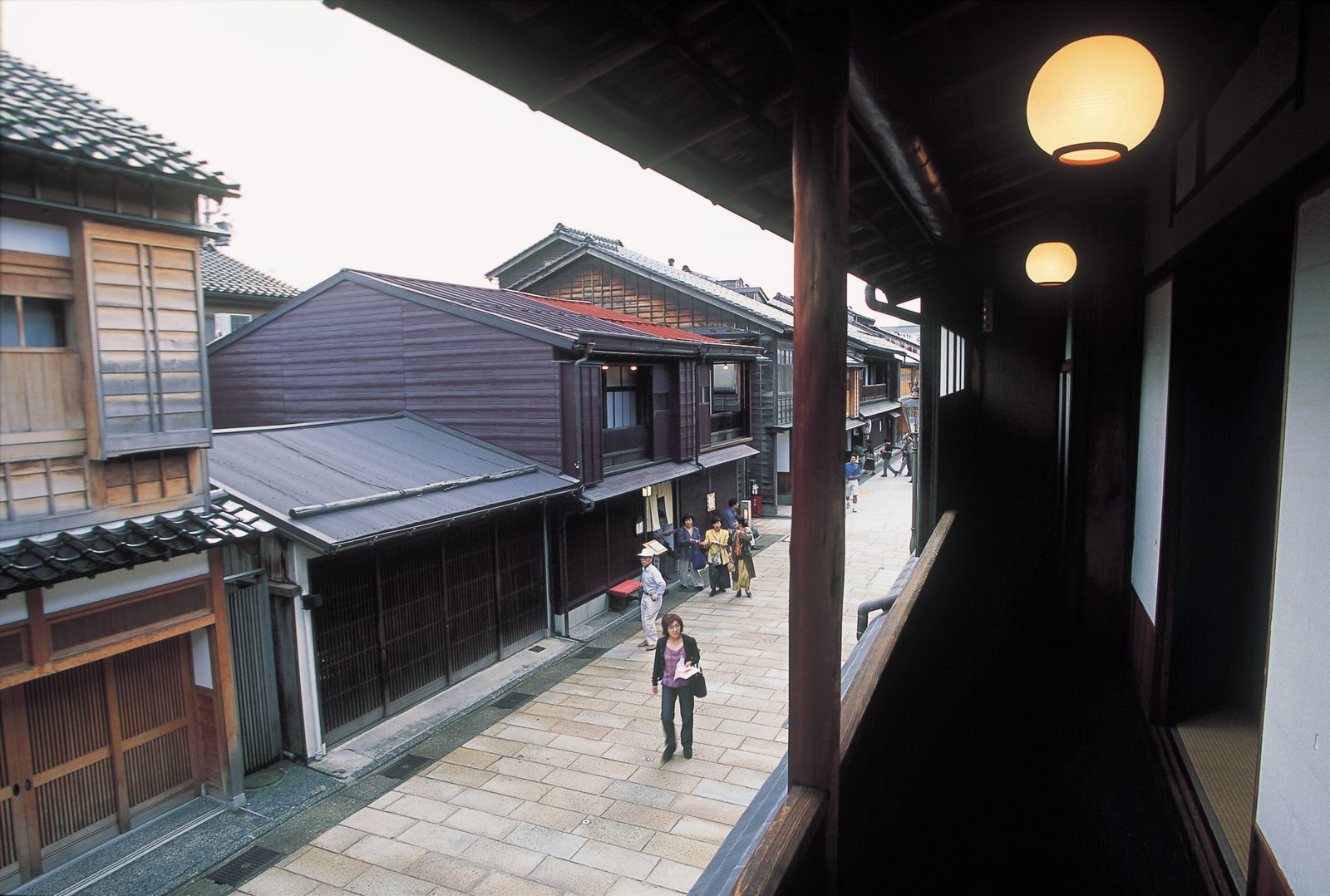A narrow lane in the Higashi Chaya geisha district at dusk, lined with dark wooden teahouses and round paper lanterns, with a few pedestrians walking past.
