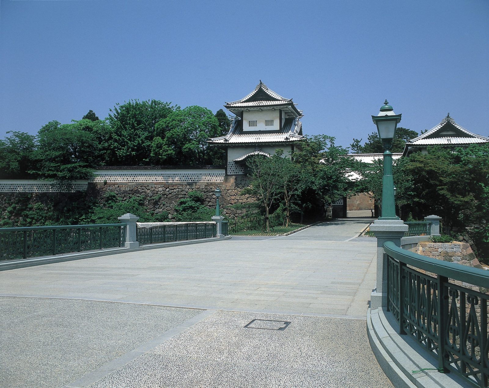 Ishikawa-mon, the turreted stone gate of Kanazawa Castle Park, seen across an empty stone bridge and plaza under a clear summer sky.