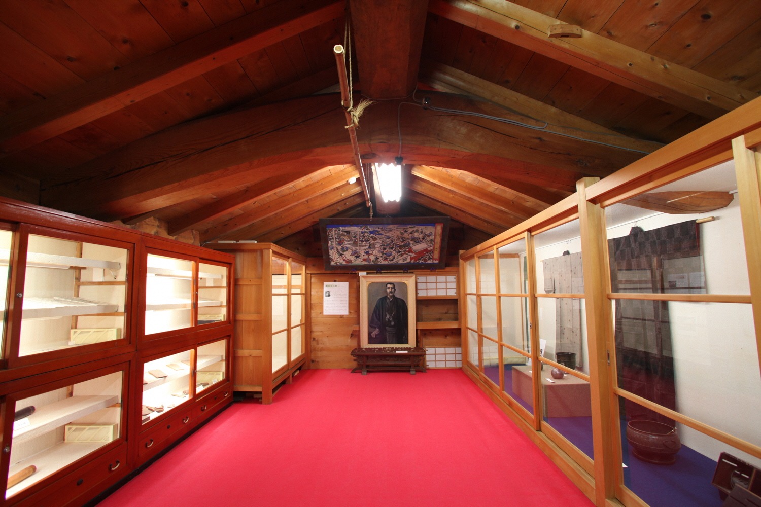 Display cases along a red-carpeted exhibition room under exposed wooden beams at the Kitamaebune no Sato Museum