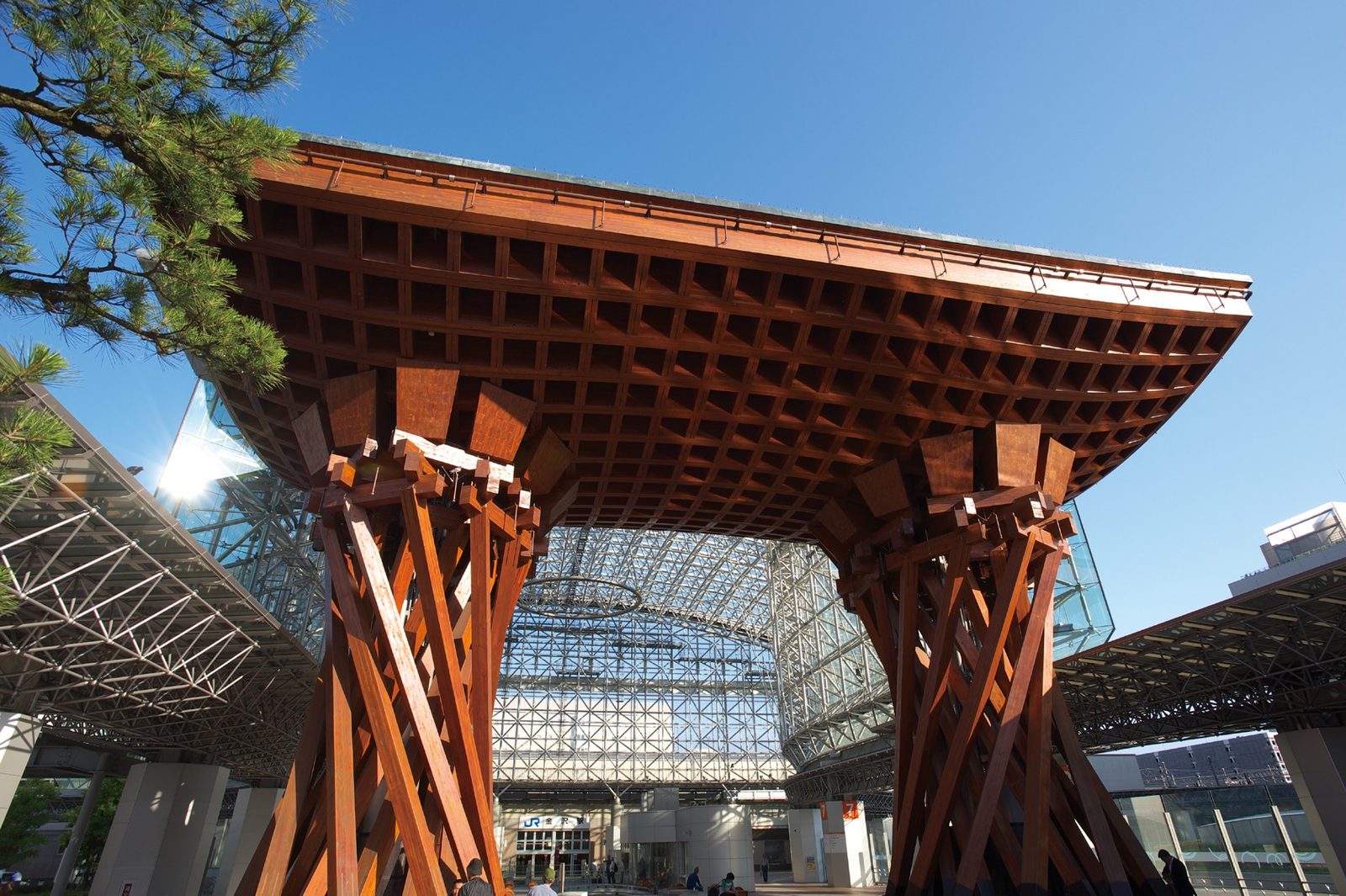 The wooden Tsuzumi-mon gate outside Kanazawa Station, its drum-shaped lattice frame and curved beams silhouetted against a clear blue sky.