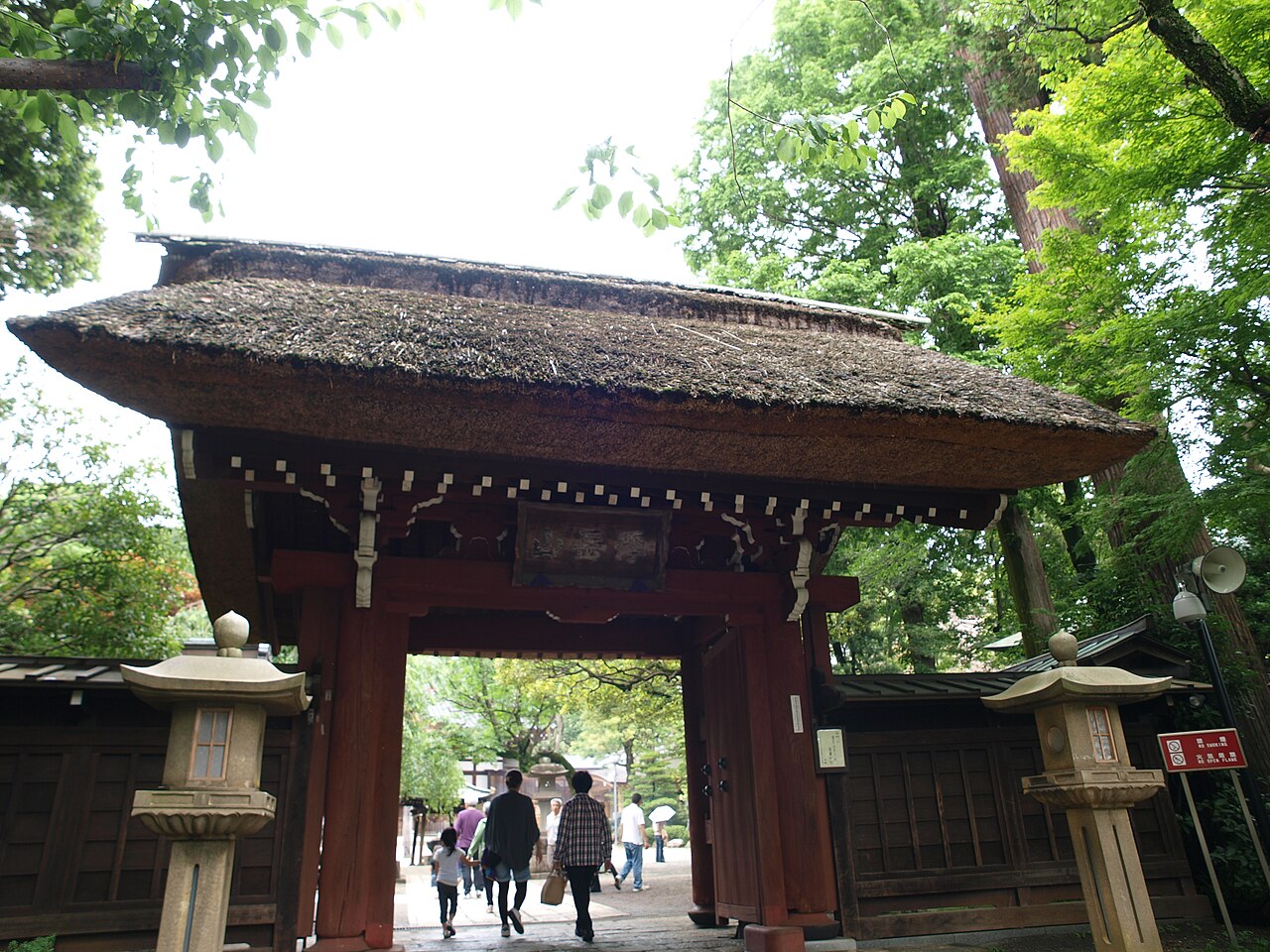 The thatched-roof mountain gate of Jindaiji Temple, with stone lanterns on either side and visitors passing beneath it.