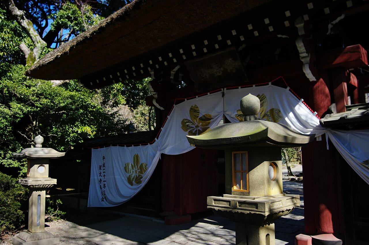 The thatched-roof gate of Jindaiji Temple in Tokyo, with stone lanterns in front and a white temple banner hanging beside it.