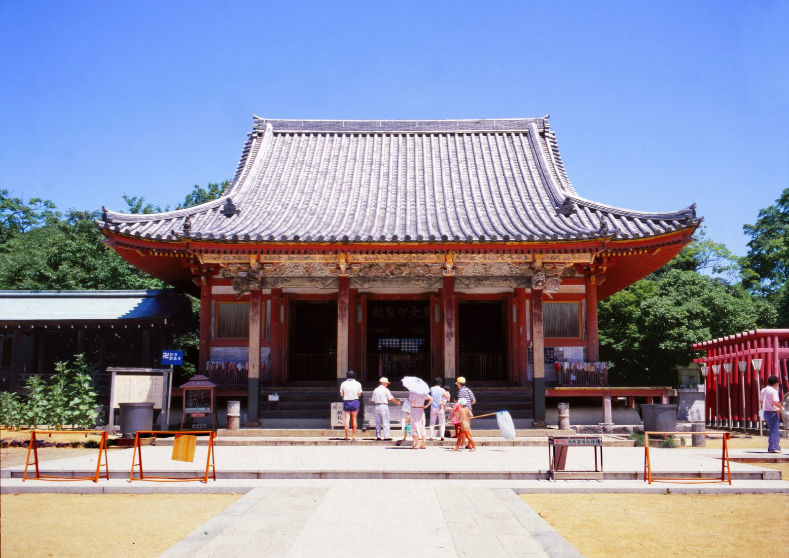 The red-pillared main hall of Yashima-ji temple with a tiled hip-and-gable roof and a few visitors on the paved forecourt under a clear sky