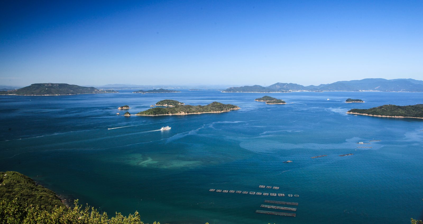 A bright blue stretch of the Seto Inland Sea dotted with small green islands and fish farm rafts, seen from a hillside viewpoint