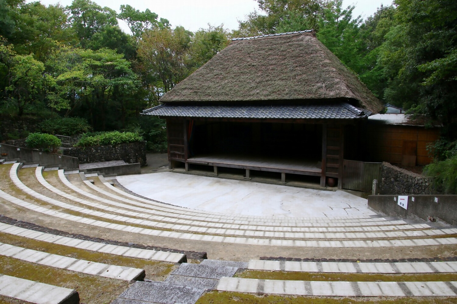 A thatched-roof open-air kabuki stage facing curved rows of stone terrace seating, ringed by trees at Shikoku Mura