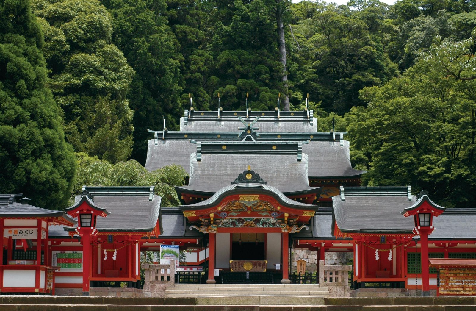 The front of Kirishima Jingu seen from the stone steps, with a red and white gate-like entrance, stacked grey roofs and a dense green forest behind.