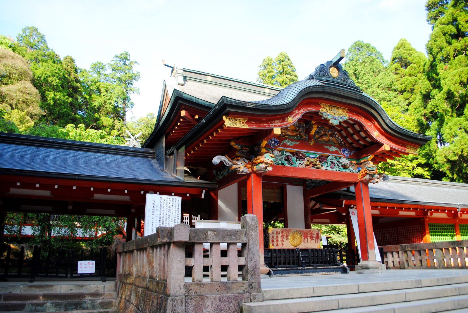 A close view of the red-lacquered front hall of Kirishima Jingu, with a curved dark roof, carved and gilded decoration, a stone balustrade and green cedar trees behind.