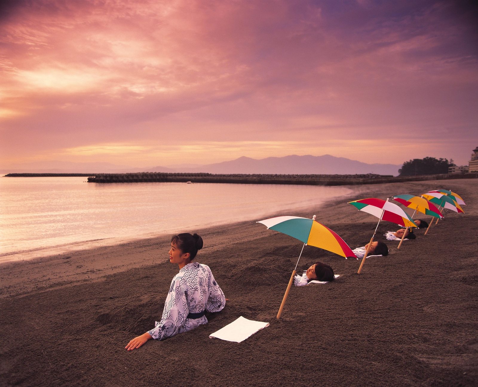 Bathers lying in dark sand under colored parasols on the Ibusuki shore at dusk, with hills across the bay