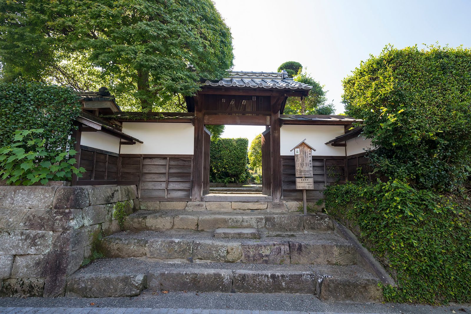 Stone steps leading up to a tiled gate set in a hedge and stone wall at a samurai house in Izumi Fumoto