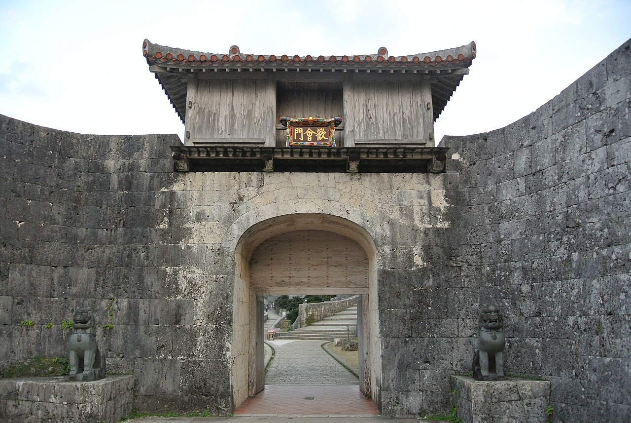 Kankaimon, a stone archway gate in the outer wall of Shuri Castle, topped by a wooden gatehouse with a tiled roof and a lacquered plaque bearing its name, flanked by two stone lion-dog (shishi) statues.