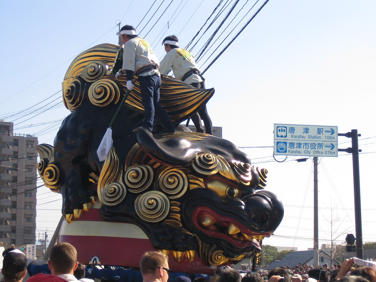 A black lion-head Karatsu Kunchi float being pulled along a street near Karatsu Station, with two riders in festival jackets on top holding the ropes
