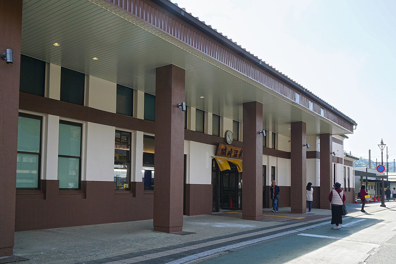 A wide two-story building with brown pillars and a tiled awning displays a yellow sign with Japanese characters and a round clock, while a few visitors gather at the entrance.