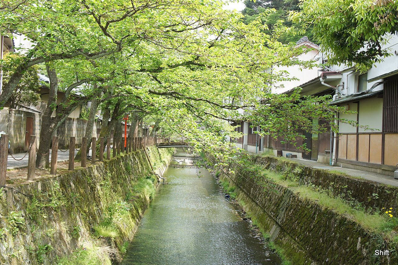 A quiet tree-lined canal winds between wooden inns and a mossy stone embankment, with a small arched bridge and green branches framing the water in daylight.