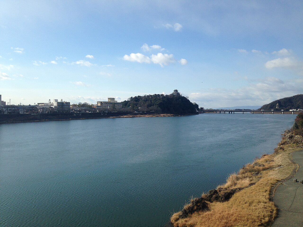 The broad Kiso River seen from the Inuyama bank, with the castle keep standing small on its wooded bluff above the far shore.
