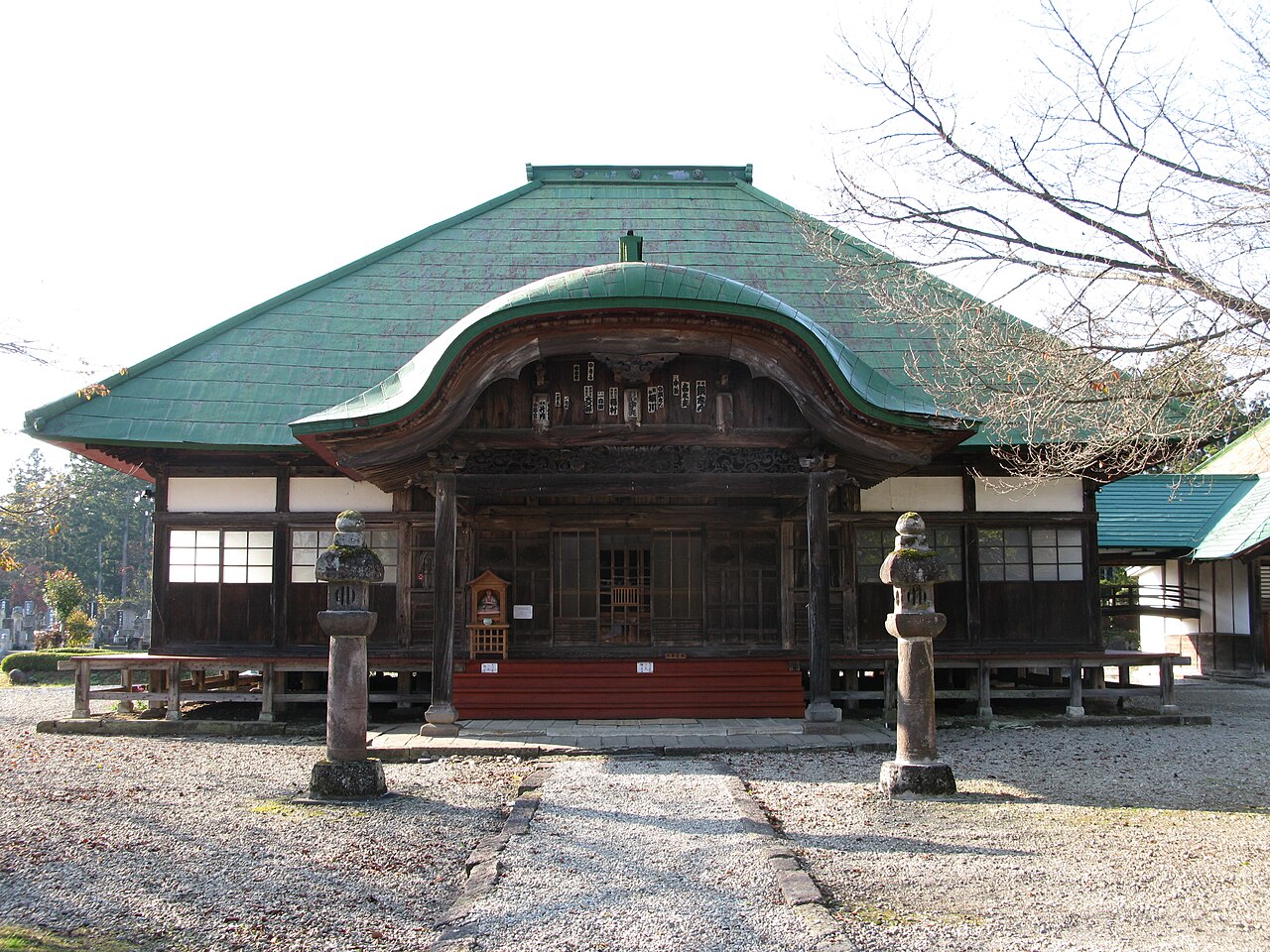 A temple hall with a green metal hip roof, a curved gable over the entrance, white plaster walls and two stone lanterns flanking a gravel approach
