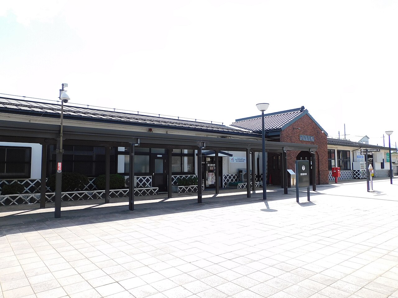 A low, dark-roofed wooden frontage with a covered walkway on slender pillars, a small red-brick gabled entrance and a wide paved forecourt under a bright sky