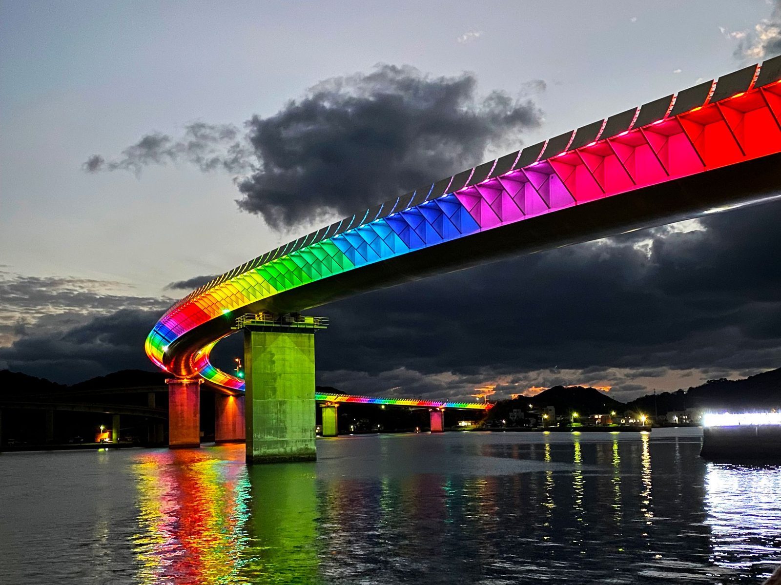 A long curving road bridge lit in rainbow colors at dusk, its concrete piers reflected in the dark harbor water under a cloudy sky