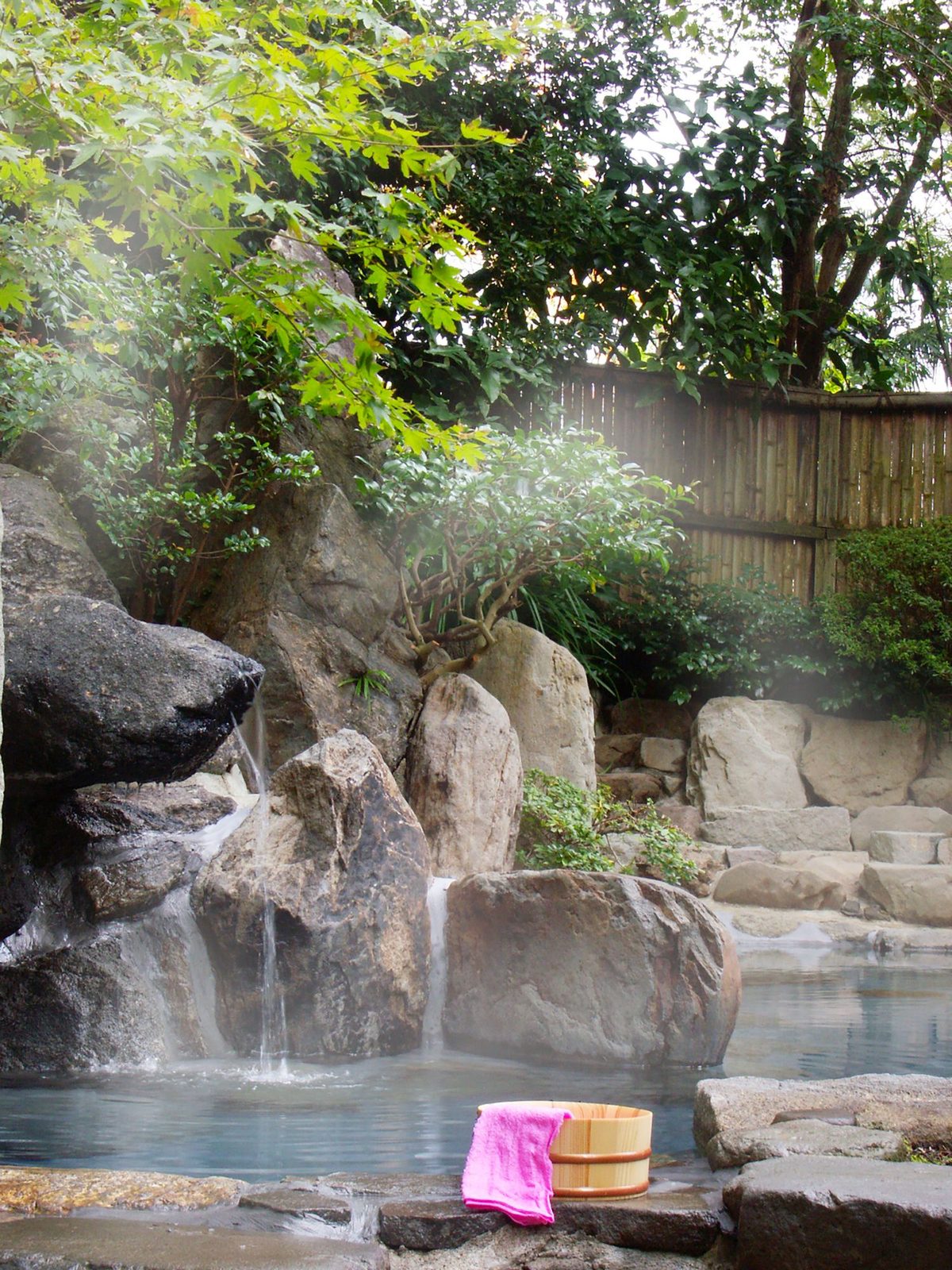 An outdoor rock-lined hot spring bath with a small waterfall trickling over boulders, a wooden bucket and pink towel at the edge, and green maple leaves above a bamboo fence.