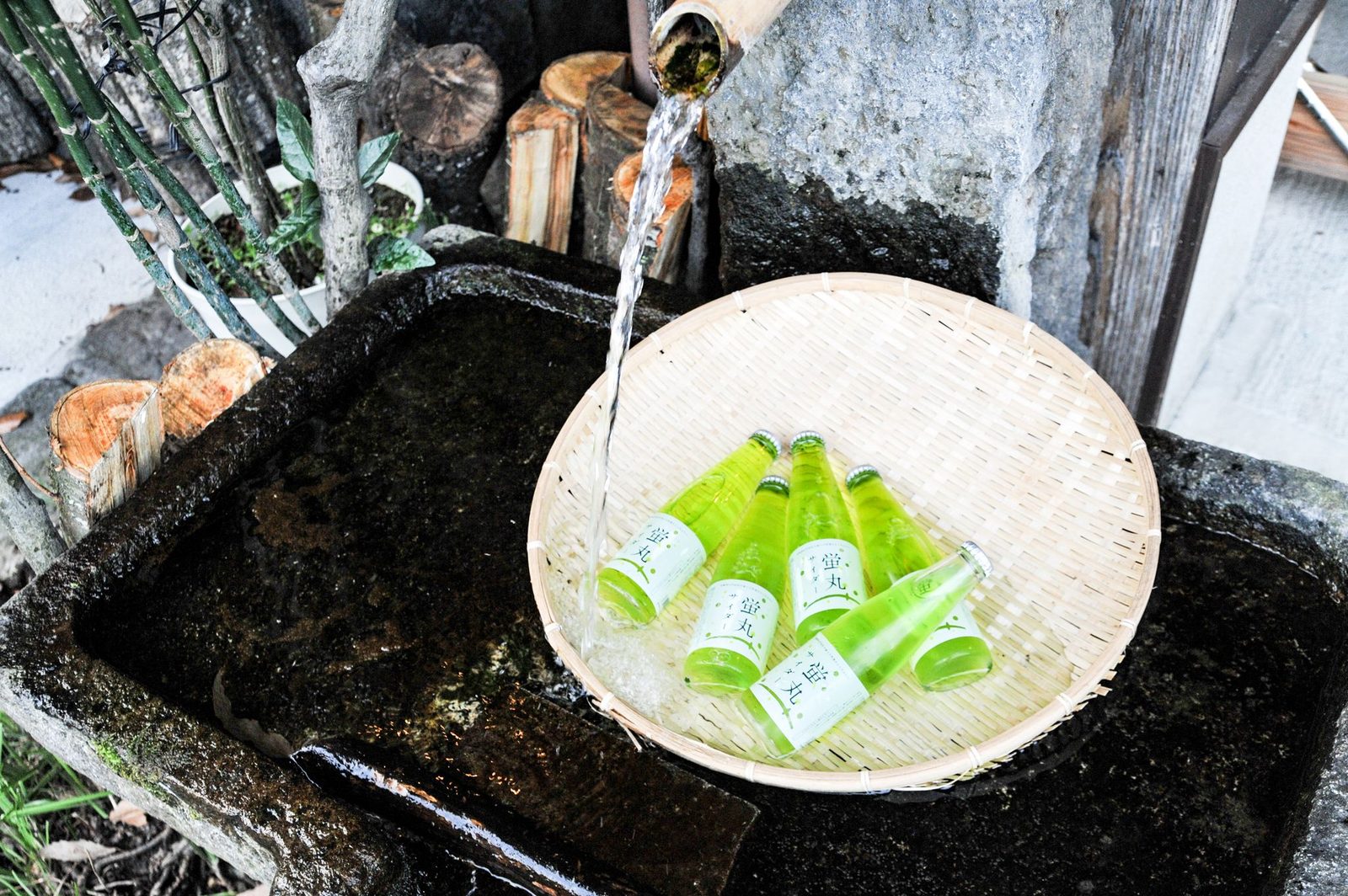 Five green cider bottles cooling in a bamboo basket under a stream of spring water running from a bamboo pipe into a mossy stone trough.