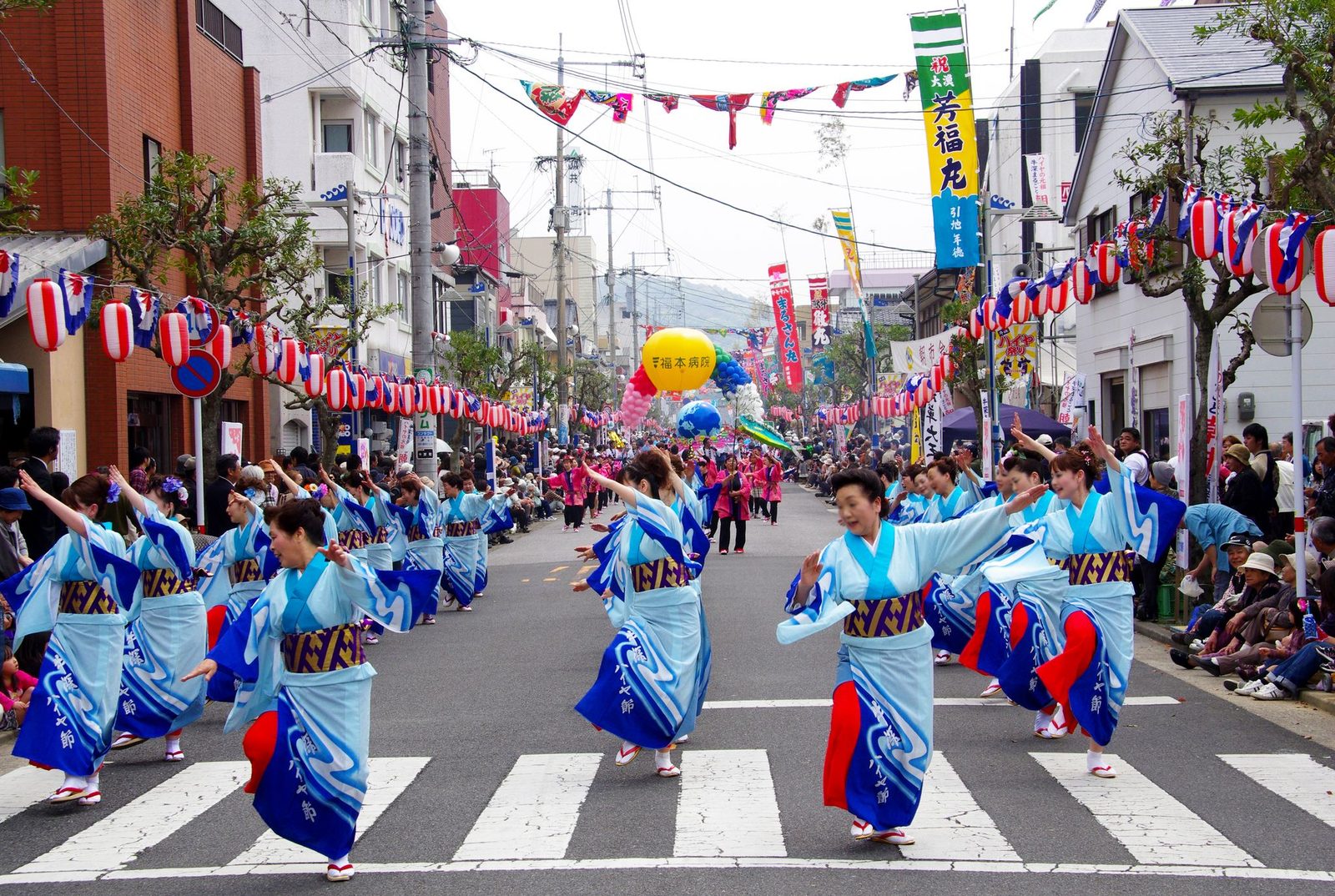 Women in pale-blue kimono with wave patterns dancing down a closed town street lined with red-and-white lanterns and festival banners, with spectators along both sides