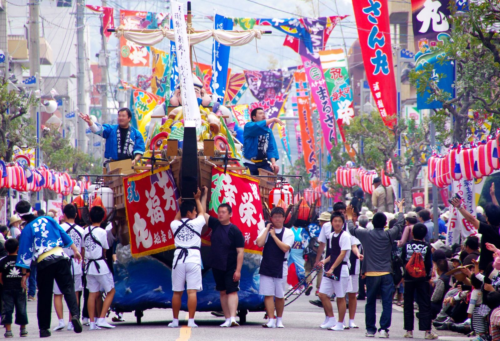 A festival float shaped like a ship carrying a large smiling figure, pulled down a street hung with colorful fishing-boat flags, with spectators on both sides