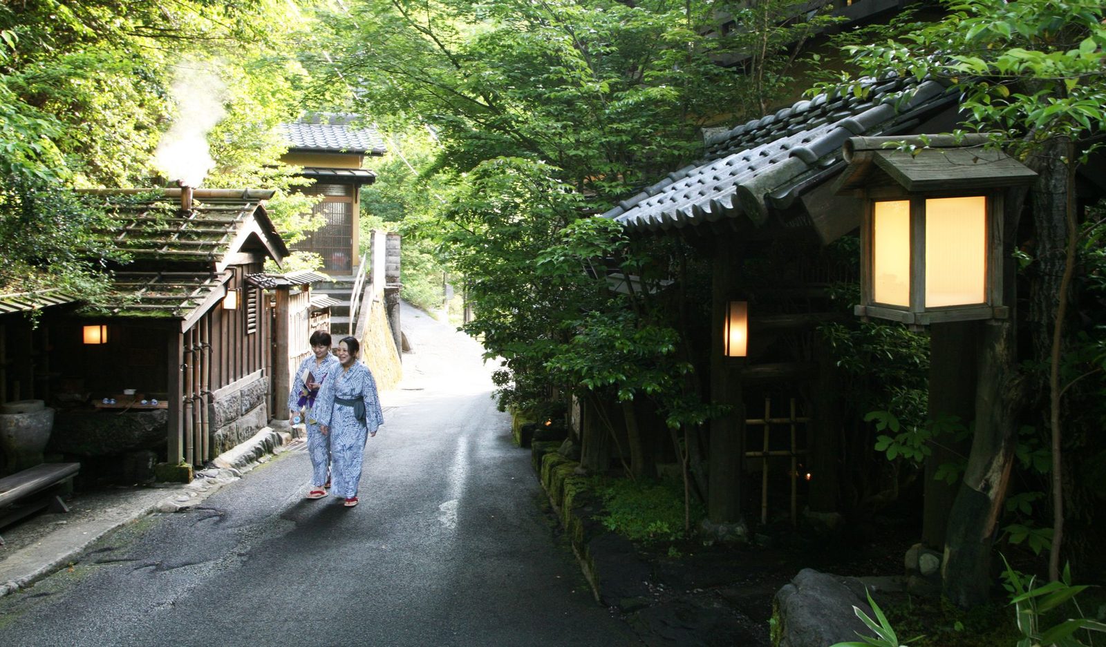 Two visitors in yukata walking along a narrow lane between dark wooden inn buildings and fresh green trees in Kurokawa Onsen
