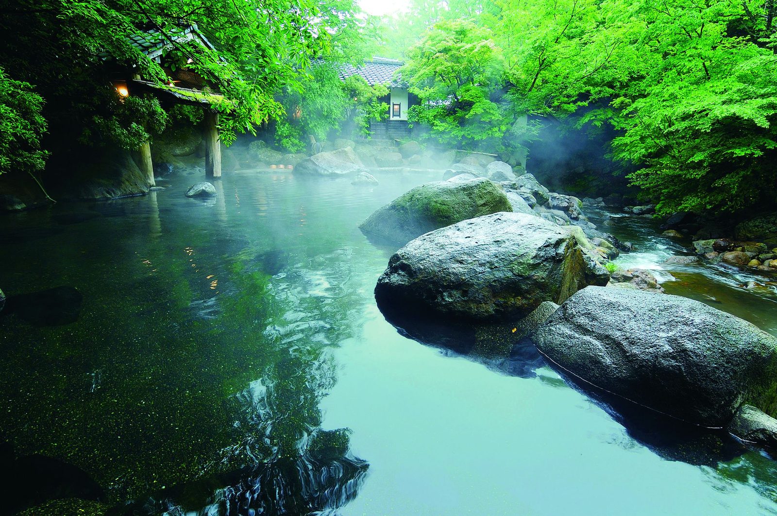 Steam rising over a riverside open-air bath framed by large rocks and green maple leaves in Kurokawa Onsen