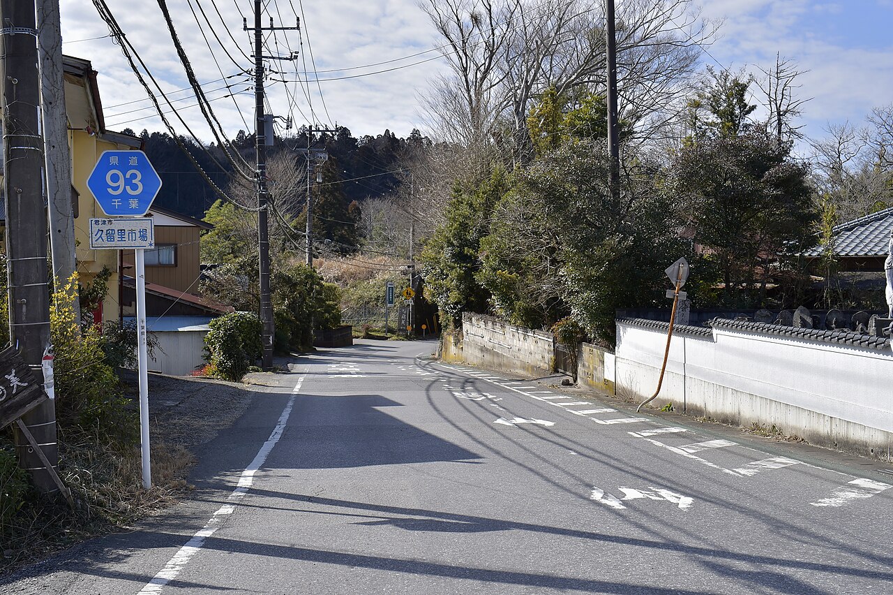A quiet road through Kururiichiba, marked by a blue prefectural route sign, leading past old houses and a walled temple toward the wooded hill where Kururi Castle stands.