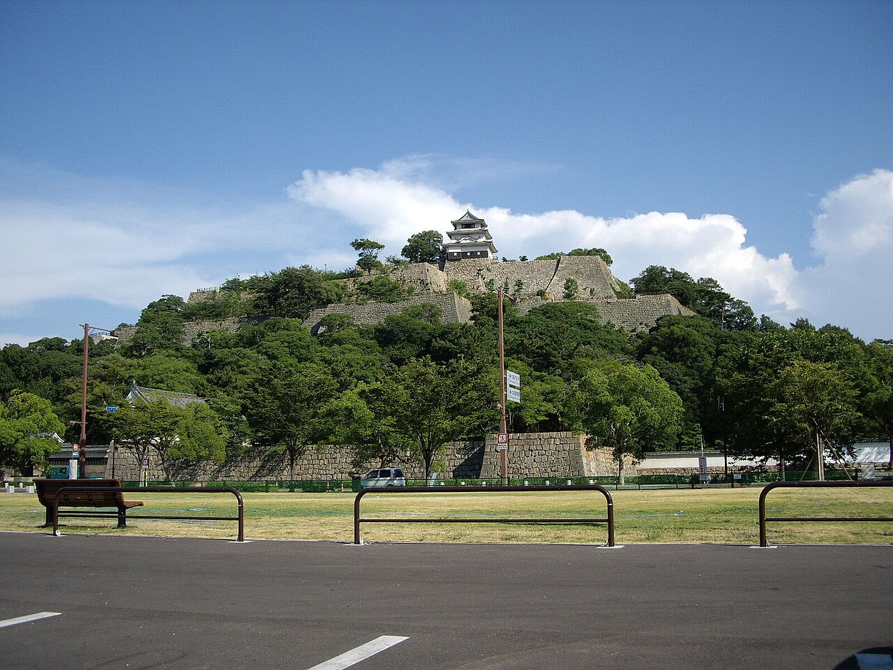 Marugame Castle seen from the park below: tiers of fitted stone ramparts climb the wooded hill, with the small three-storey white keep standing at the top.