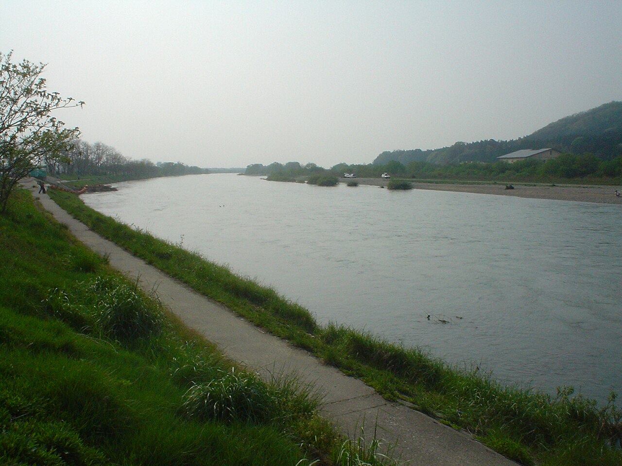 The broad, slow Miomote River seen from a grassy embankment, with a paved walking path along the near bank and low wooded hills on the far side under a pale sky.