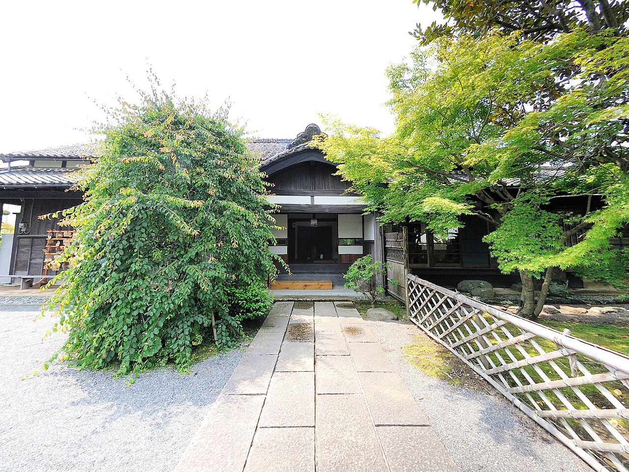 A low wooden house with a tiled roof set behind a paved stone path, shrubs and maple trees, in the old centre of Mishima.