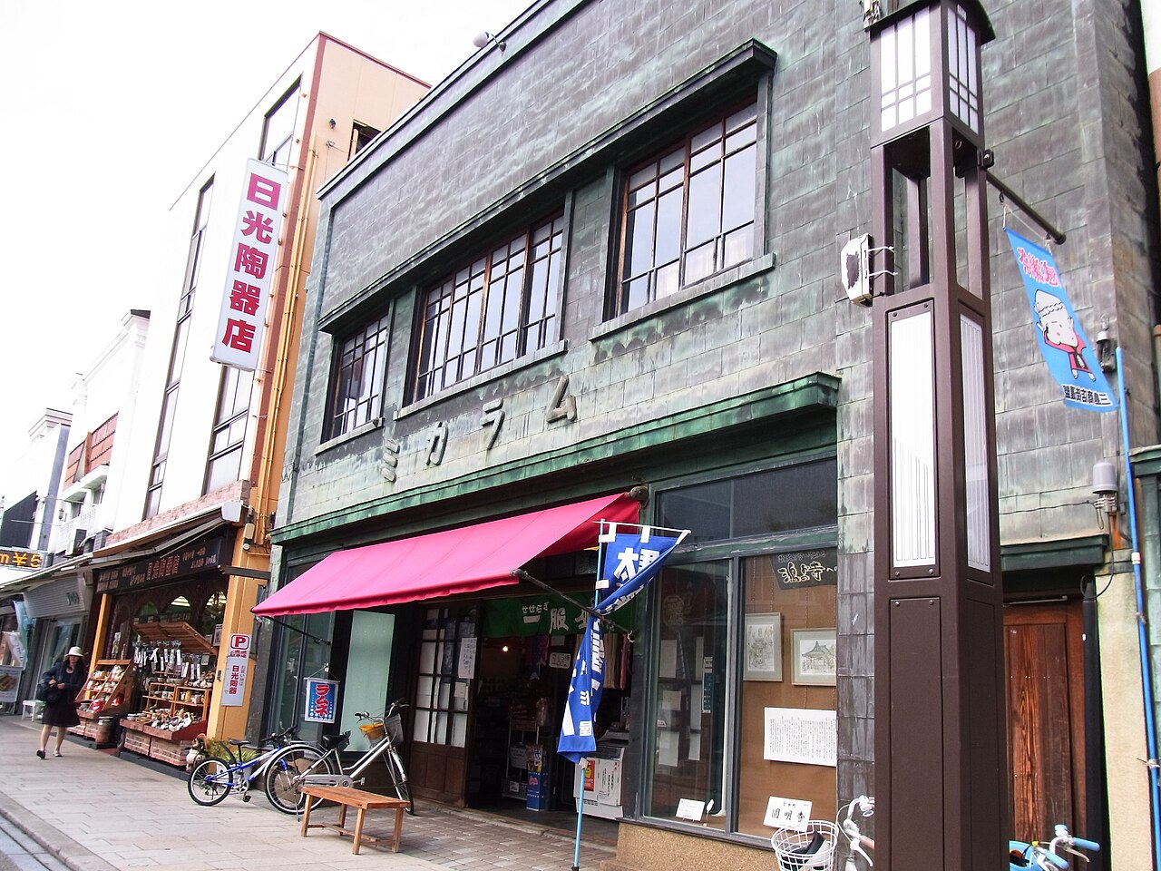A two-storey shop front clad in weathered green copper sheeting, standing among smaller shops on a street in Mishima.