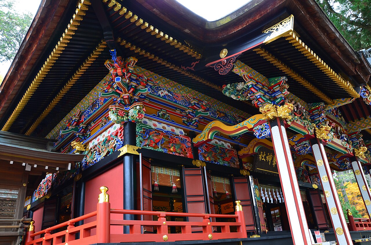 A shrine building at Mitsumine with deeply carved eaves painted in red, gold, blue and green, its vermilion railing and dark pillars set against cedar forest.