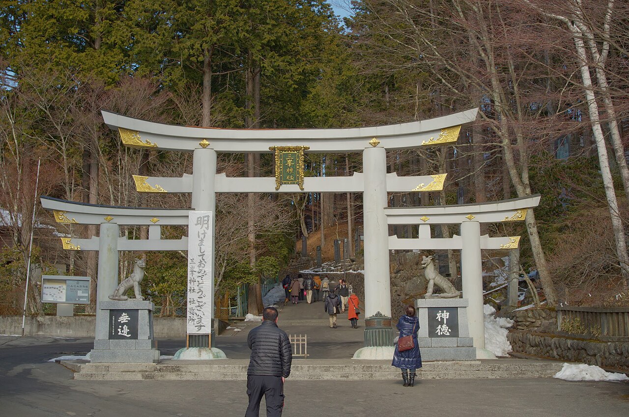 The unusual three-part torii gate at the entrance to Mitsumine Shrine, flanked by seated stone wolf guardians instead of the usual lion-dogs, with visitors walking up the cedar-lined approach beyond it and patches of snow at the roadside.