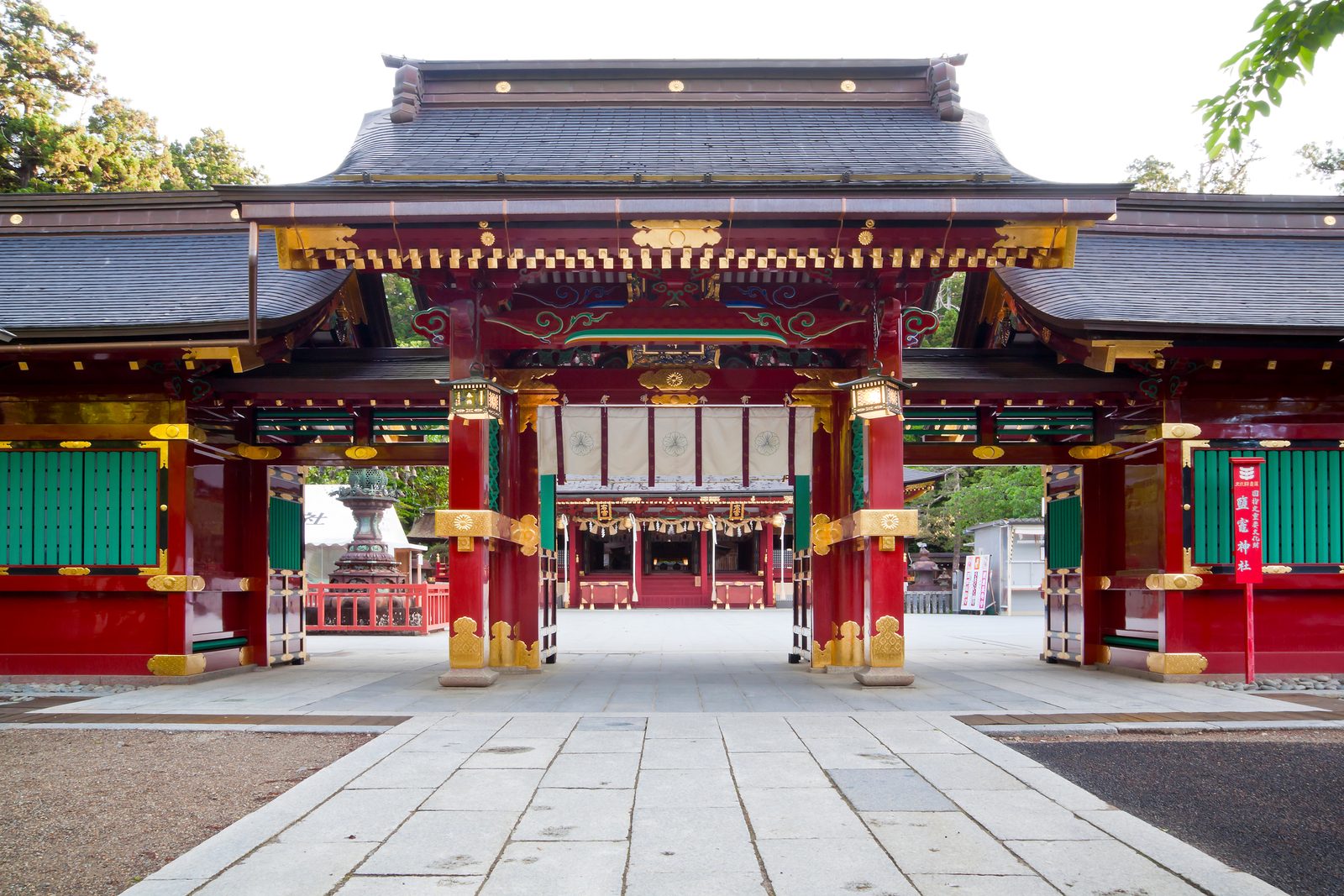 The vermilion gate of Shiogama Shrine with green side wings, framed by pine and maple trees beyond the stone-paved approach