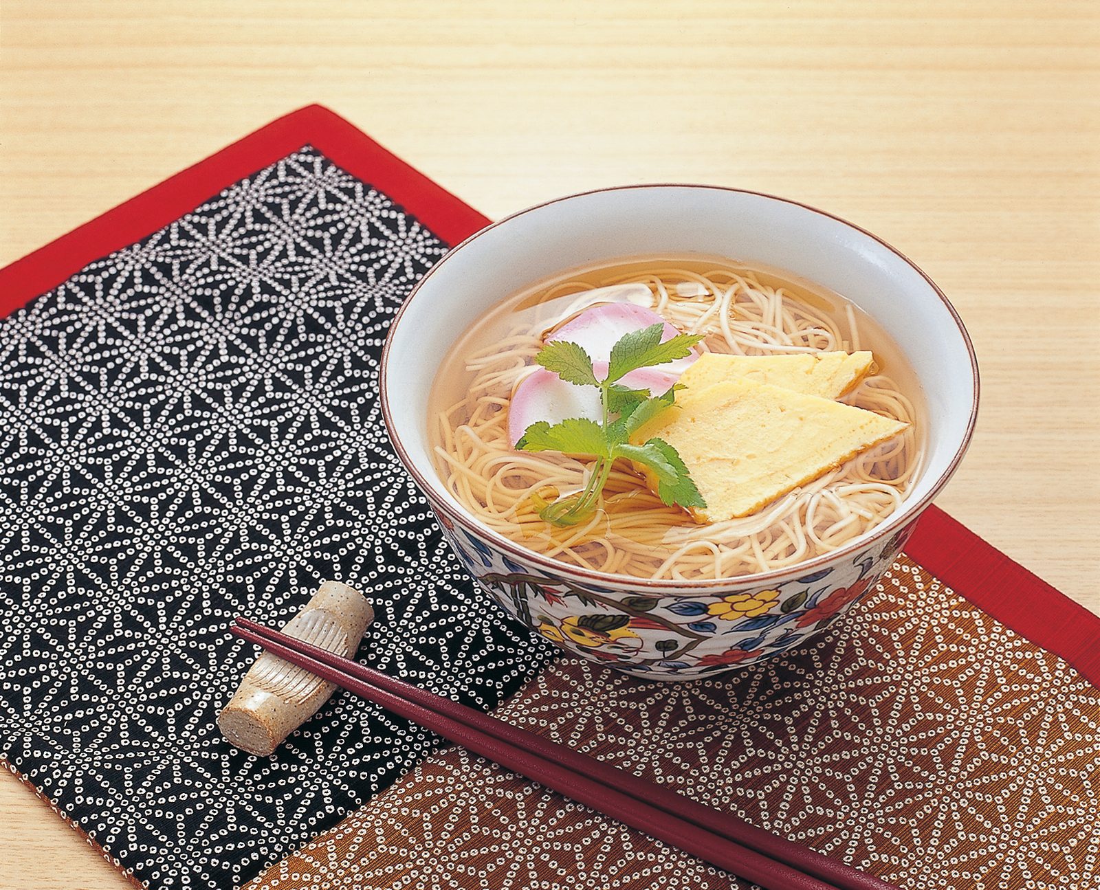 A bowl of white Shiroishi umen noodles with fish cake and a leaf garnish, served on a patterned mat with chopsticks.