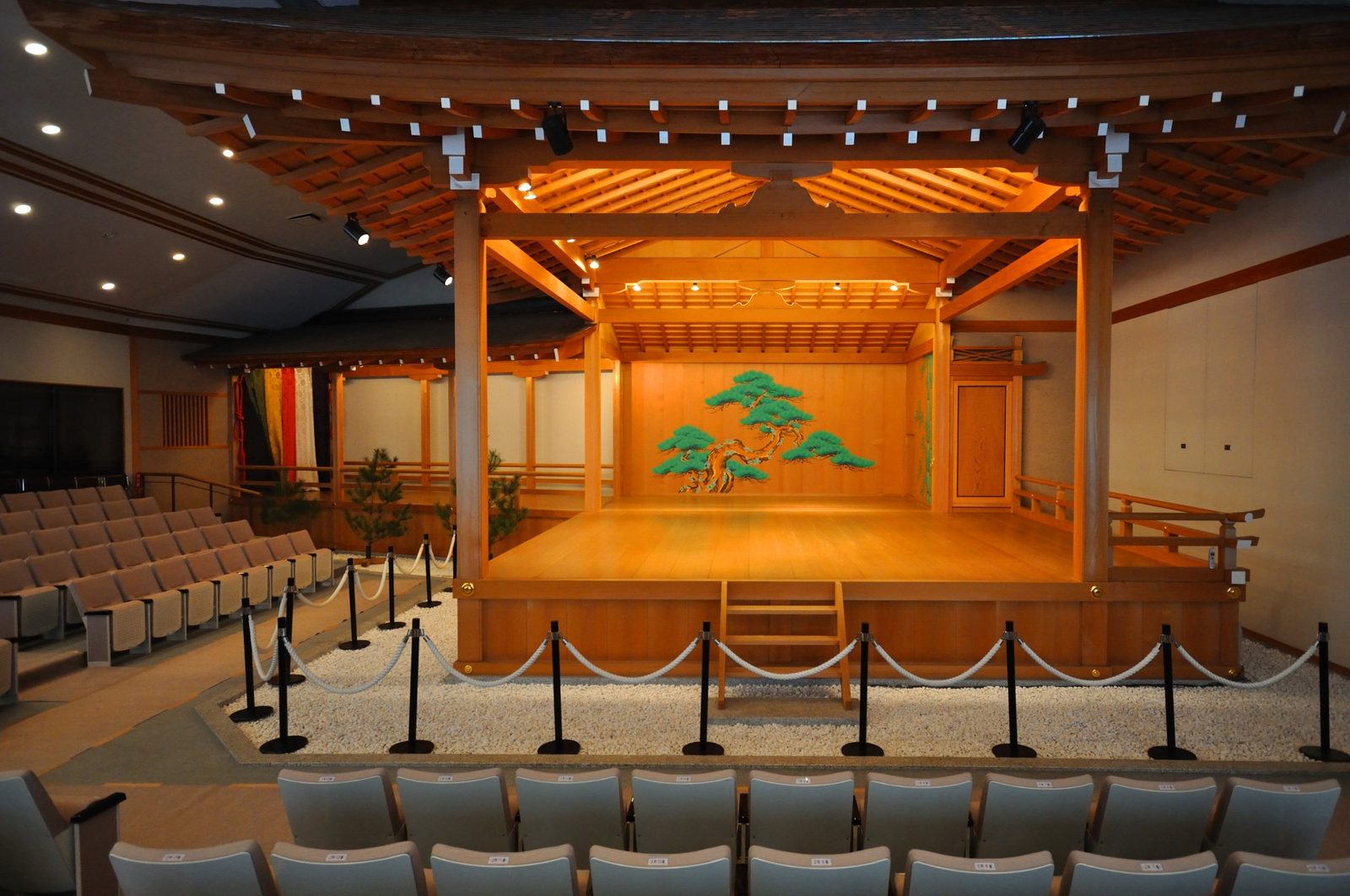 The wooden Noh stage with a pine painted on its back wall, facing rows of seats in an indoor theater.