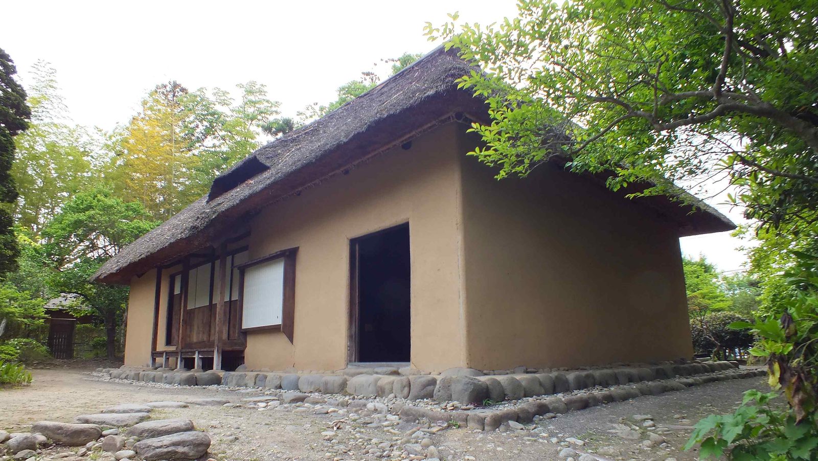 A thatched-roof samurai residence with plastered earthen walls, surrounded by trees.