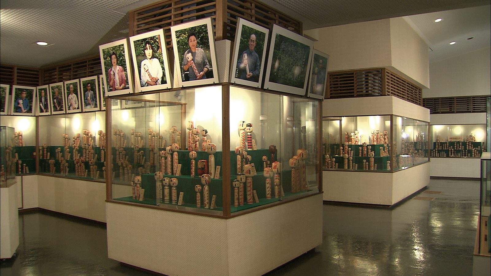Wide view of a museum gallery with glass cases full of painted wooden kokeshi dolls beneath framed portrait photographs of kokeshi makers.