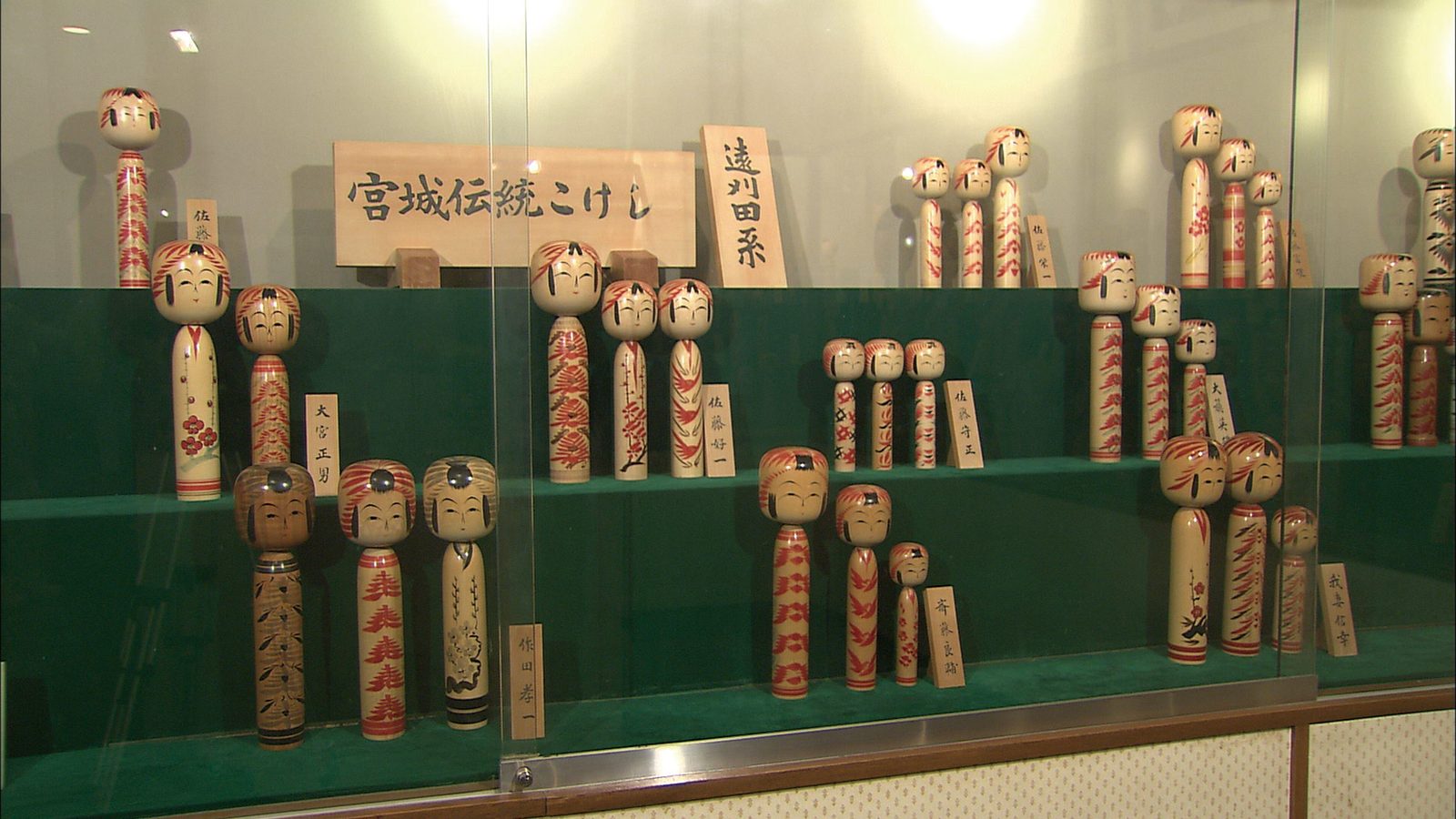 Glass display case of traditional kokeshi dolls with red floral and feather patterns beside wooden signs naming the Miyagi kokeshi tradition and the Togatta lineage.