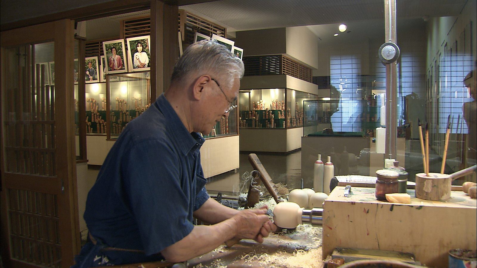 A grey-haired craftsman in a blue work shirt shapes the rounded head of an unpainted kokeshi on a lathe, wood shavings across the bench.