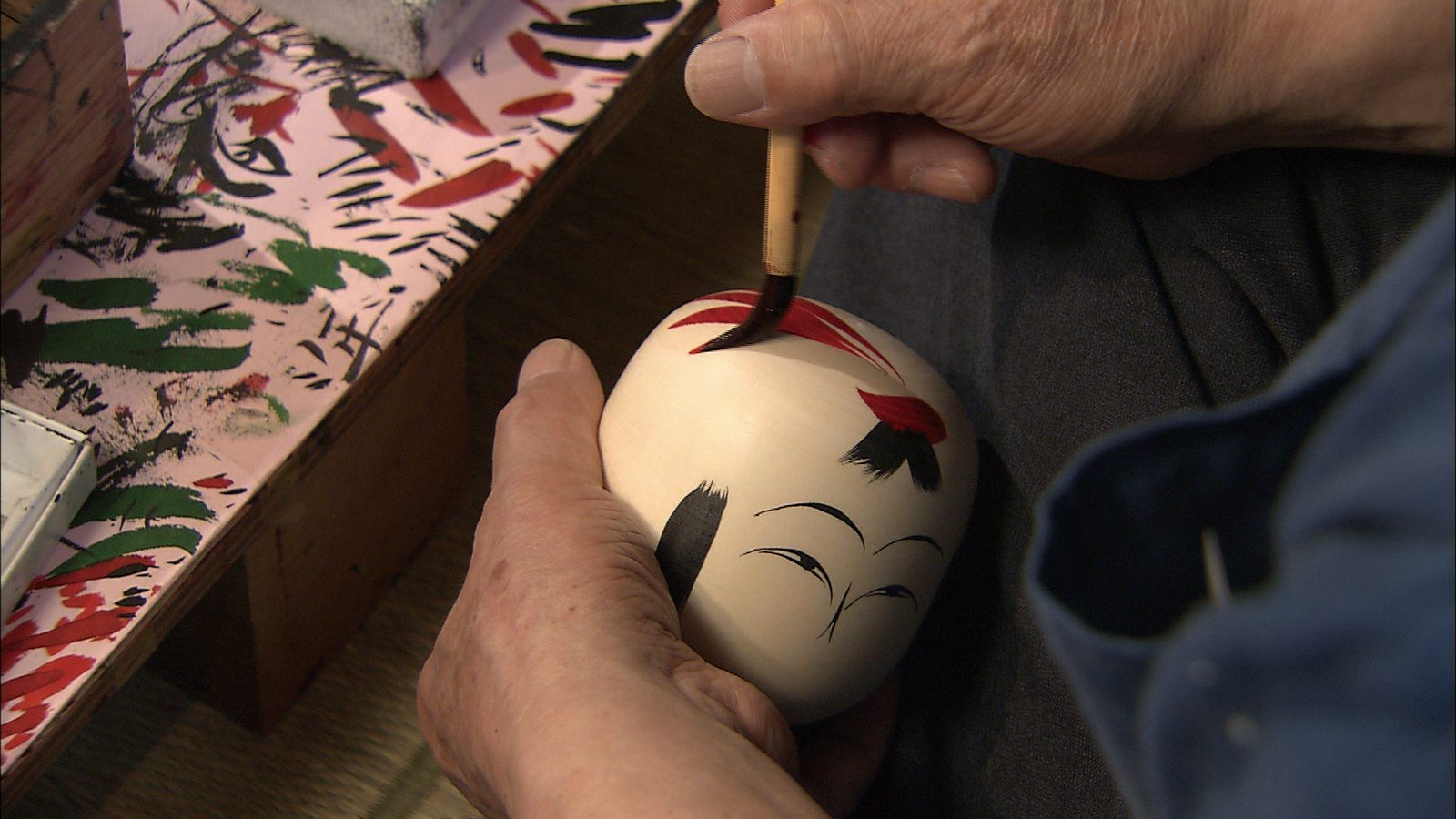 Close-up of a craftsman's hands brushing red and black strokes onto the head of a wooden kokeshi doll.