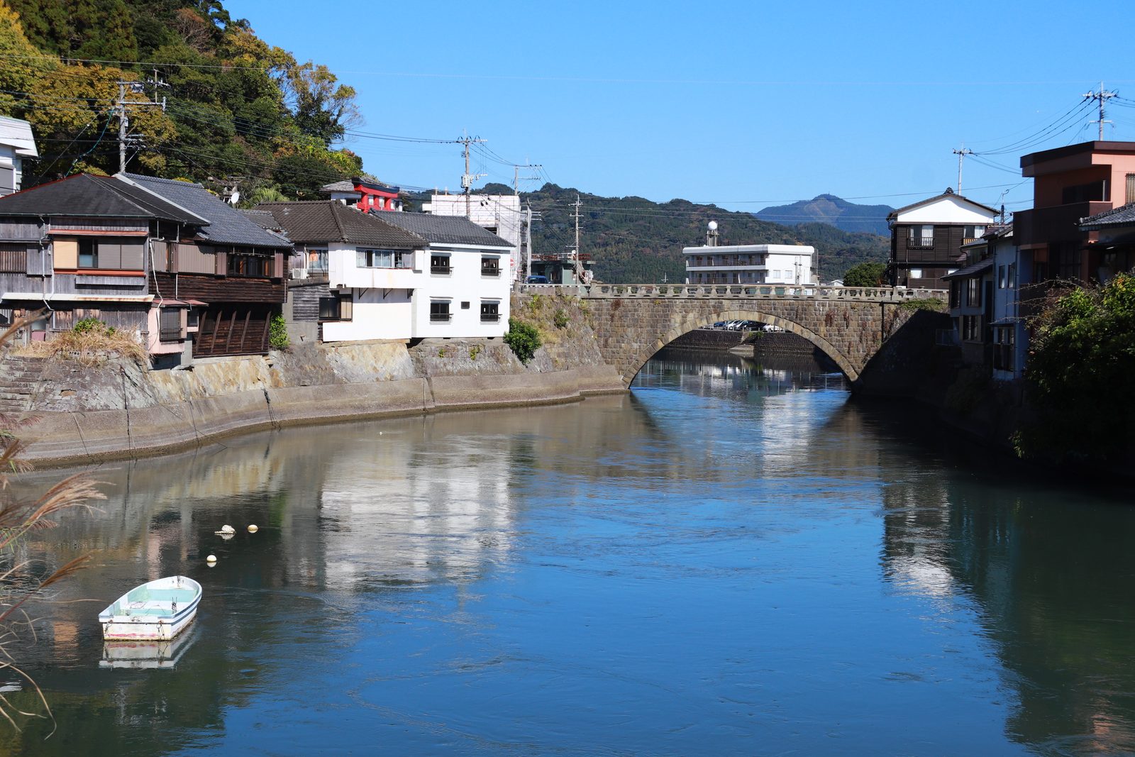 A stone arch bridge over a calm canal reflecting old wooden and white houses, with green hills behind.