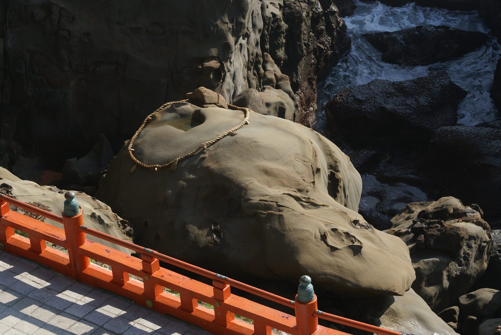 A rounded boulder ringed with a sacred straw rope beside a red railing, on rocks above the sea.