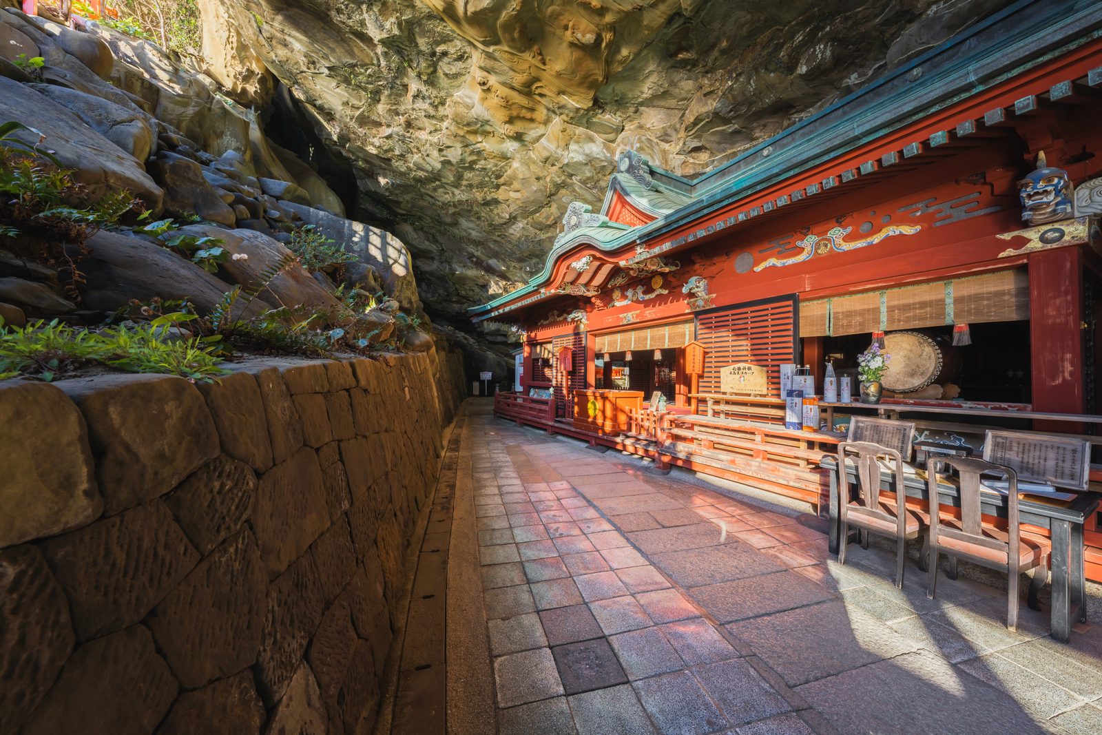 A vermilion shrine hall built inside a sea cave, with a stone-paved path running beneath the overhanging rock.