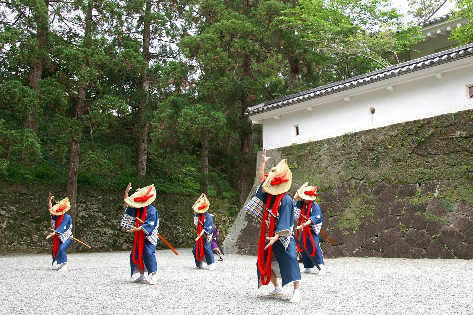 Performers in indigo kimono and straw hats with red padded neck wraps parade across a gravel courtyard beside a white plastered stone wall at Obi Castle.