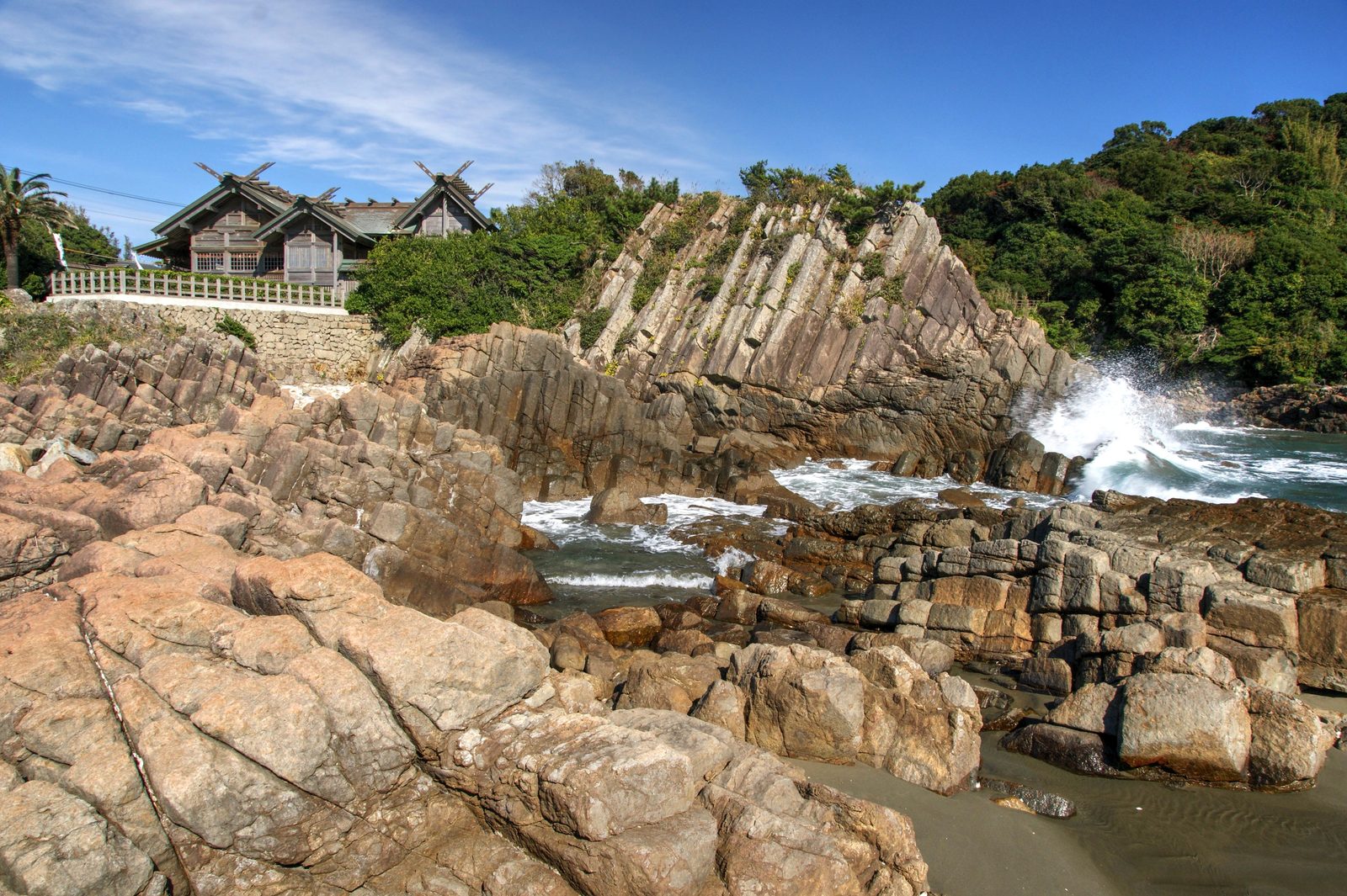 A shrine hall standing on a rocky shore above the sea, with columnar-jointed rock beside it