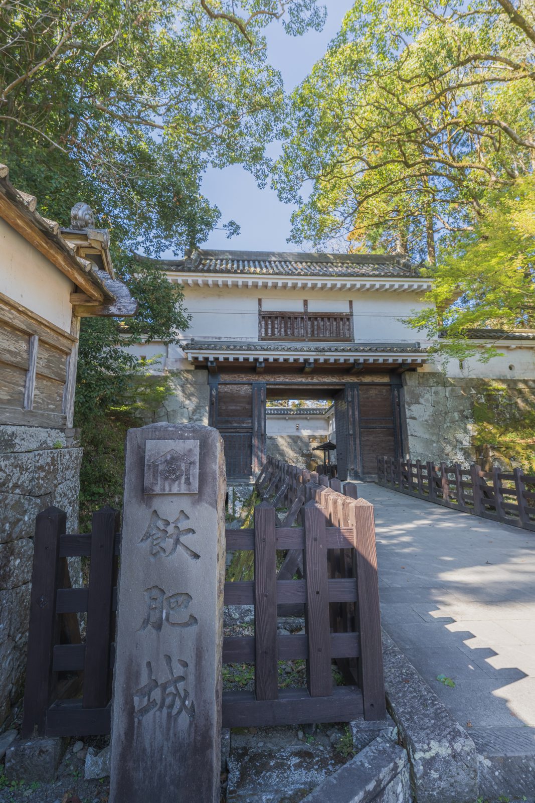 A carved stone marker for Obi Castle stands beside a two-story wooden and white-plastered castle gate, reached by a stone bridge lined with tall trees.