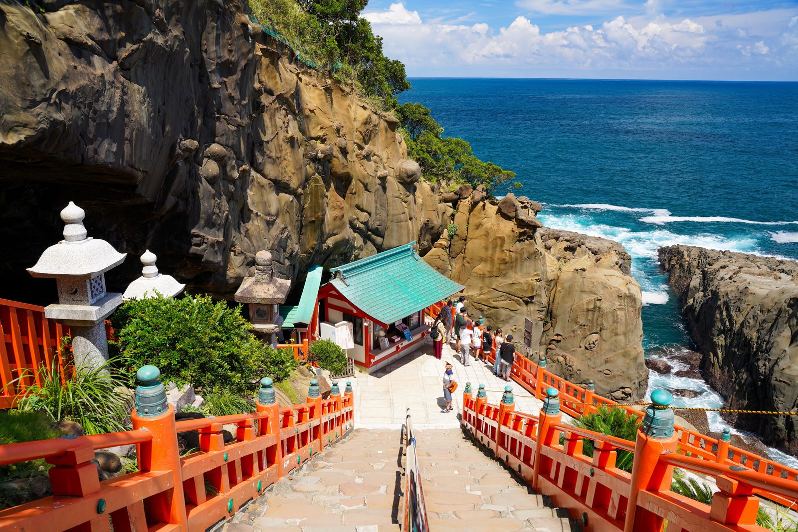 A red-railed stone stairway descending toward a small teal-roofed shrine building on a rocky coast, with the blue sea beyond.