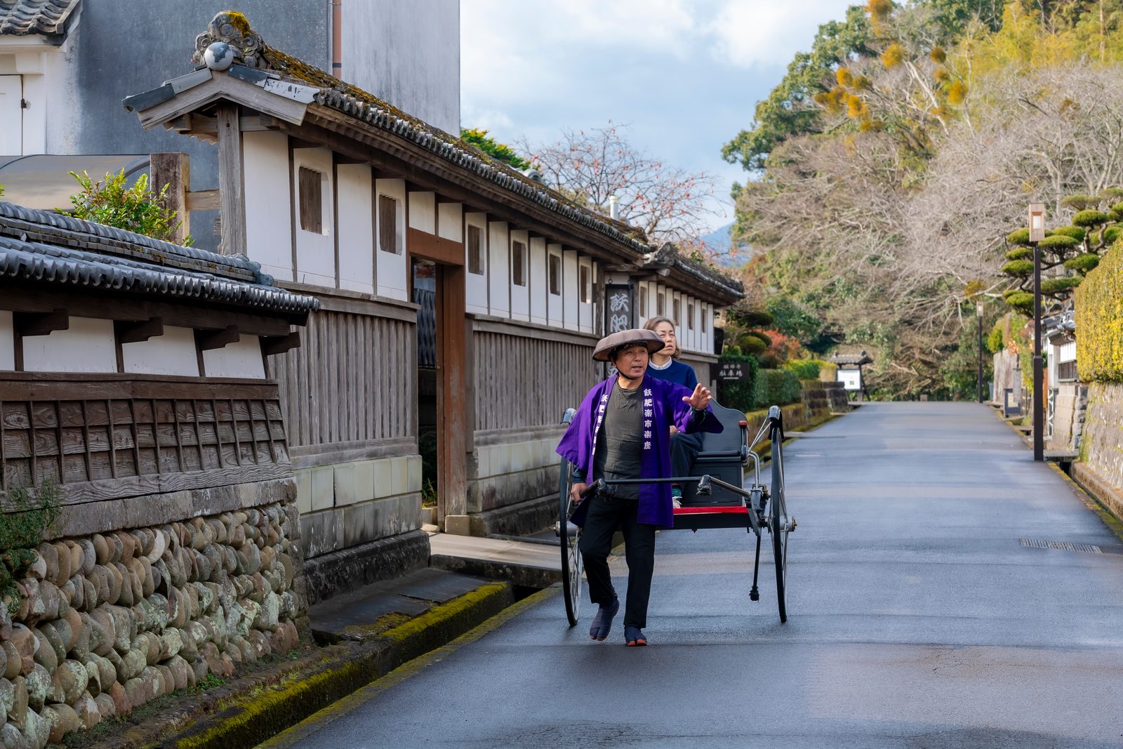 A rickshaw puller in a purple happi coat pulls a passenger past white plastered walls and stone foundations on a quiet street in Obi's old castle town.